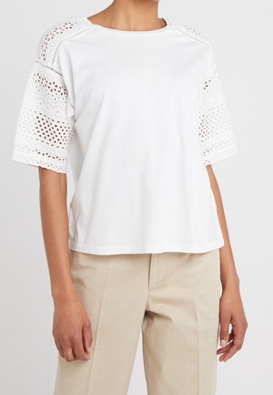 Top blanco de manga corta con patrones de encaje troquelados en las mangas, combinado con pantalones beige, mostrando la parte superior del cuerpo y los brazos.