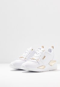 Weiße Sportschuhe mit einem strukturierten Obermaterial, goldenen Hardware-Akzenten und eckigen, metallischen Fersendetails. Mit Schnürsenkeln und Logo-Branding.