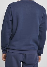 Persona con un sudadera azul marino sin dibujos y pantalones a juego, vista por la espalda, de pie frente a un fondo claro.
