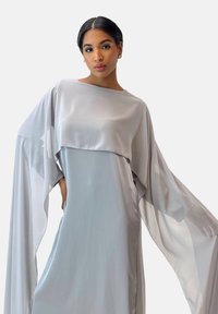 Robe grise fluide en tissu léger avec de larges manches évasées et une fente haute. Douce brillance avec un design en couches au niveau du corsage.