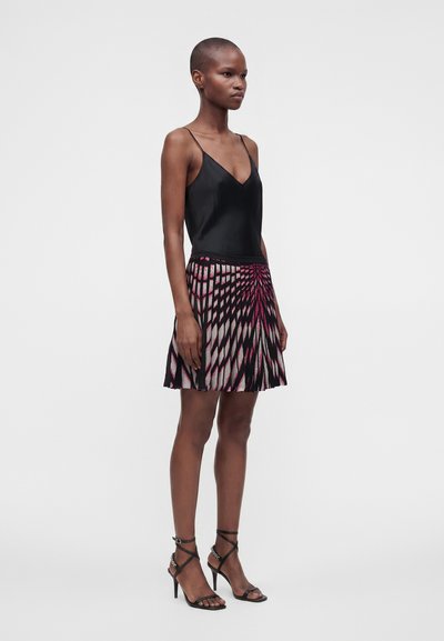 Femme portant un top en satin noir, une jupe plissée à motifs rouges et beiges avec un design géométrique, et des sandales noires à talons hauts avec des lanières.