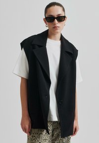 Svart ärmlös blazer med slag, strukturerad design och en enkel knappstängning, parat med en vit t-shirt och mönstrade byxor.