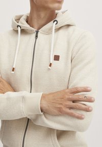 Beige hettegenser med glidelås laget av strukturt stoff, med kontrasterende snører og en liten brun logo-patch. Har ribbestrikkede mansjetter og nederkant.