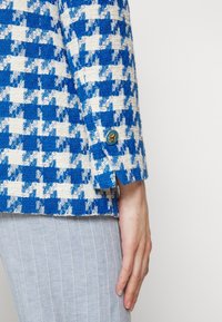 Manche de veste à motif pied-de-poule bleu et blanc avec un bouton décoratif sur une jupe côtelée gris clair, main reposant de manière décontractée sur le côté.