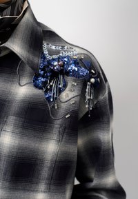 Geruite shirt in marineblauw en grijs met textuurstof. Kenmerkt zich door een decoratieve blauwe pailletten bloemaccent en parelversieringen op de schouder.