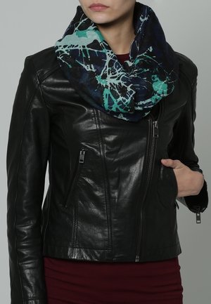 Femme portant une veste en cuir noire sur un haut bordeaux et une écharpe infinie à motifs bleu-vert autour du cou.