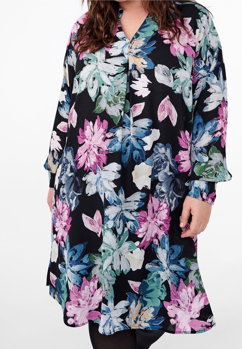 Femme portant une robe noire à manches longues avec de grands imprimés floraux en rose, bleu, vert et blanc.