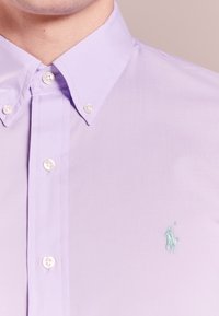 Ljus lavendelfärgad skjorta med knappar och spetsig krage, med vita knappar och en liten blå broderad logotyp på bröstet. Slät tygtextur.