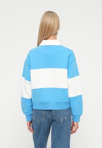 Mujer rubia vistiendo un sudadera de bloques de color azul y blanco y jeans azules, de pie frente a un fondo blanco con la espalda a la cámara.