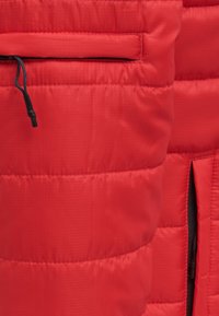 Gros plan sur un tissu de veste matelassée rouge avec des coutures horizontales et un cordon de fermeture éclair noir sur une poche.
