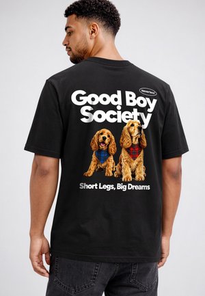 Mann trägt schwarzes T-Shirt mit dem Text "Good Boy Society" und zwei Spanielhunde mit blauen und roten Schals, die darunter sitzen.