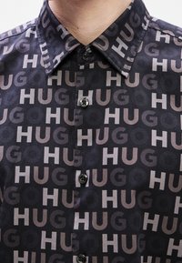 Chemise noire boutonnée avec un motif répétitif de grandes lettres "HUG" en gris et tons plus foncés sur tout le tissu.