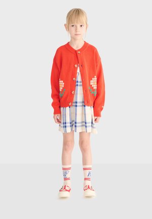 Ragazza giovane che indossa un cardigan rosso con bottoni e motivi floreali, un vestito a quadri blu e bianco, calzini decorati e scarpe rosse, in piedi su uno sfondo semplice.