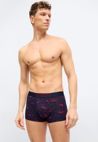 Άνδρας χωρίς πουκάμισο, φορώντας σκούρο μπλε μποξέρ με κόκκινες καρδιές και σχέδια κροκοδείλου, στέκεται μπροστά σε απλό φωτεινό φόντο.