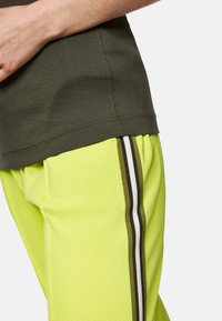 Olive green, ribbed t-shirt paired with bright yellow, textured joggers featuring brown and white side stripes. Close-up of the waist area.