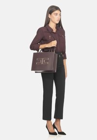 Borsa a tote in pelle bordeaux con forma strutturata, dettaglio del logo in tono oro e doppi manici superiori, abbinata a pantaloni neri e tacchi.