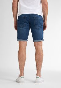 Shorts in denim blu con risvolti, dotati di due tasche posteriori e una vestibilità casual, abbinati a sneakers bianche da ginnastica.