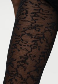 Πόδι διακοσμημένο με μαύρα διαφανή καλσόν με σχέδιο λουλουδάτου αμπελιού πάνω σε σκούρο δέρμα σε λευκό φόντο.