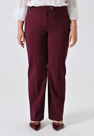 Donna che indossa pantaloni burgundy su misura e décolleté coordinati, in piedi su uno sfondo bianco neutro.