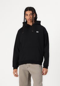 Schwarzer Hoodie mit Fronttasche, aus weichem Stoff gefertigt. Er verfügt über ein kleines Logotag auf der Brust und gerippte Bündchen und Saum.
