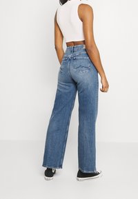 Jeans de mezclilla azul de cintura alta con pierna recta, que cuentan con bolsillos traseros y un detalle de costura sutil, combinados con zapatillas negras.