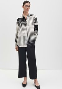 Schwarz-weiß gestreiftes Hemd mit Knöpfen, langen Ärmeln und lockerem Sitz, kombiniert mit schwarzen, weit geschnittenen Hosen und spitz zulaufenden Schuhen.