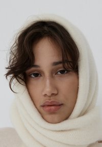 Biały dziergany szalik owinięty wokół twarzy, o miękkiej fakturze i subtelnych odcieniach, na jasnym tle.