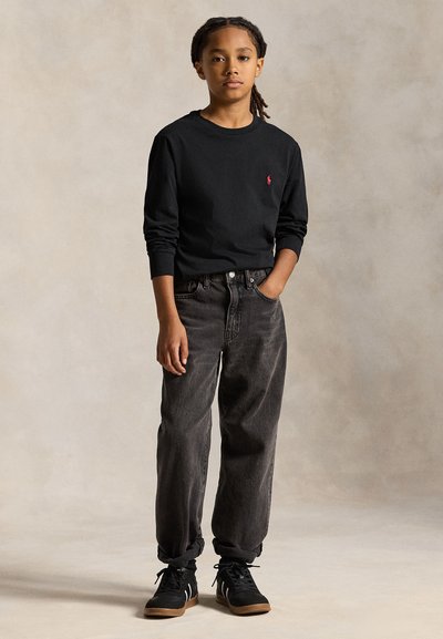 Copil cu părul împletit stă cu o mână în buzunar, purtând un tricou negru cu mâneci lungi, blugi largi închis la culoare și adidași negri.