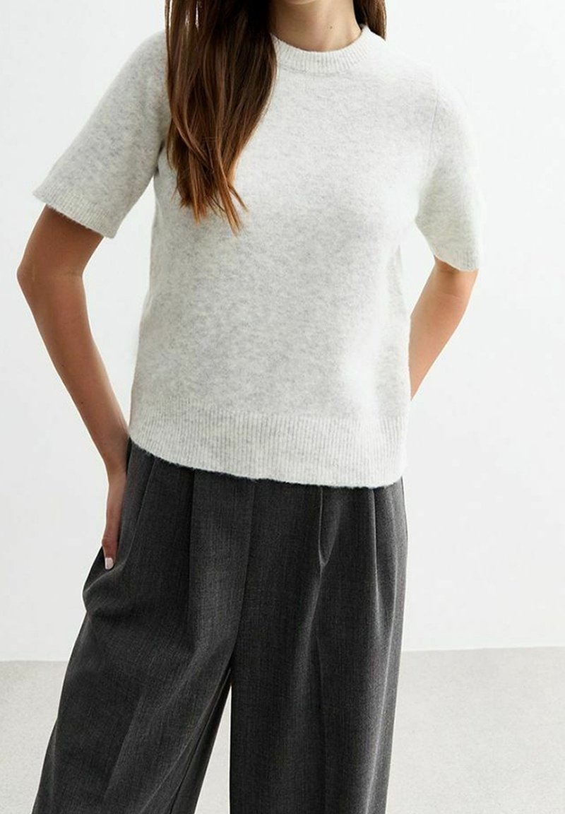 Femme portant un pull en maille gris clair à manches courtes et un pantalon plissé ample gris foncé, les mains dans les poches.