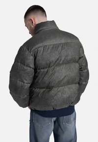 Sötét szürke puffermellény, texturált felülettel, magas gallérral és bordázott tengerészkék mandzsettákkal. A hátsó nézeten egy steppelt dizájn és tömör forma látható.