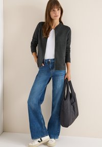 Giacca grigia con zip sopra una maglietta bianca, abbinata a jeans blu a gamba larga. Tiene una borsa tote nera. Sneakers con un dettaglio del logo.
