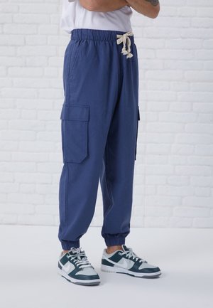 Persoon in blauwe cargojoggingbroek met elastische boorden en een witte koord, gecombineerd met witte en donkergroene Nike sneakers.