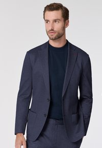 Blazer blu navy con texture, design aderente, chiusura con un bottone e due tasche frontali. Indossato sopra una maglietta scura a girocollo.