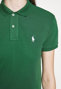 Grön polo skjorta i strukturerad tyg, med krage, tre knappslå och en liten vit broderad logotyp på bröstet.