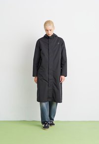 Svart vanntett frakk med høy krage og lange ermer, har glidelås foran og minimalistisk design. Båret over blå jeans og sko.