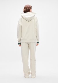 Light beige hoodie with a large hood and drop shoulders, paired with matching pants. Cuffs feature green and white stripes. Smooth texture.