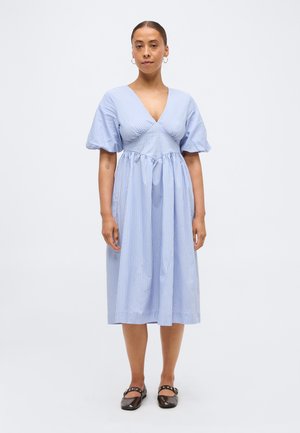 Nainen seisoo ja hänellä on päällään vaaleansininen, lyhythihainen V-kaula-aukkoinen midimekko, jossa on pystysuorat valkoiset raidat, sekä mustat tasapohjaiset kengät, joissa on remmit.