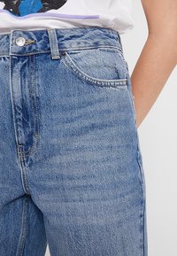 Vaaleansiniset farkut korkealla vyötäröllä, jossa on klassinen viiden taskun muotoilu, hillitty kulutusjälki ja kontrastiväriset ompeleet.