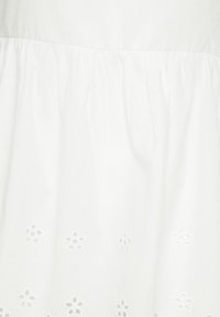 Tissu blanc avec une couture froncée et des motifs de broderie en œillets floraux le long du bord inférieur.