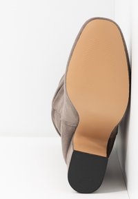 Semelle d'une bottine en daim taupe avec une semelle extérieure lisse de couleur marron clair et un talon noir texturé, positionnée debout sur un fond blanc.