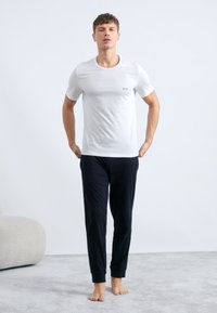 White cotton t-shirt with a small logo, paired with black relaxed-fit pants. Model stands barefoot on a light carpeted surface.