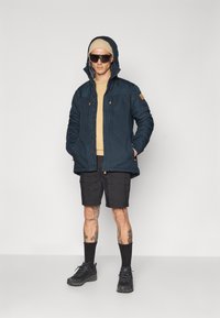 Navy padded jacket with a hood, tan sweatshirt underneath, and black cargo shorts. Accessorized with black sunglasses and dark shoes.