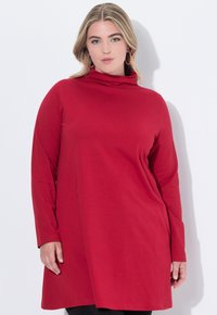 Donna con capelli biondi che indossa un abito tunica rosso a maniche lunghe e grandi orecchini a cerchio colorati, in piedi di fronte a una parete bianca.