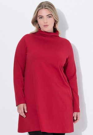 Vrouw met blond haar die een lange mouw rode tuniekjurk en grote kleurrijke hoepeloorbellen draagt, staande tegen een witte muur.