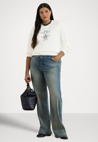 Kobieta z długimi ciemnymi włosami ubrana w biały sweter w warkocze, wyblakłe niebieskie jeansy, czarne buty, trzyma czarną torebkę, stoi na białym tle.