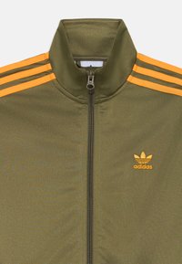Maslinasto zelena Adidas trenirka s narančastim prugama, zatvaranje na patentni zatvarač, visoki ovratnik i izvezeni logo na lijevoj strani prsnog koša. Glatka tekstura.