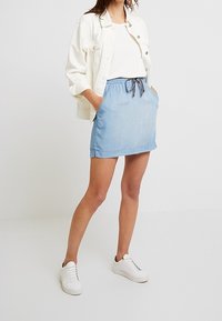 Gonna di jeans azzurro chiaro con vita regolabile, tasche frontali e finitura orlo; abbinata a una maglietta bianca e a una giacca crema. Sneakers bianche.