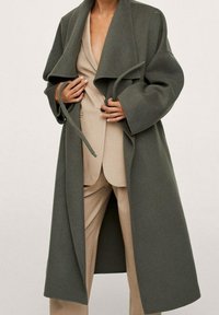 Lange, oversized jas van donkergroen wol, met brede revers, een strikceintuur en grote mouwen. Draagt over een beige pak. Soepele textuur.