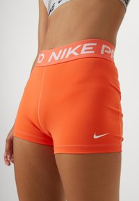 Pomarańczowe spodenki Nike Pro wykonane z elastycznego materiału, z szerokim paskiem w talii ozdobionym białym napisem "NIKE PRO" oraz białym logo Nike swoosh.