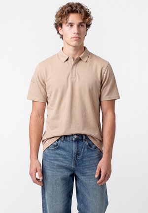 Junger Mann mit lockigem Haar, der ein beiges kurzärmeliges Poloshirt und blaue Jeans trägt, steht vor einem schlichten weißen Hintergrund.